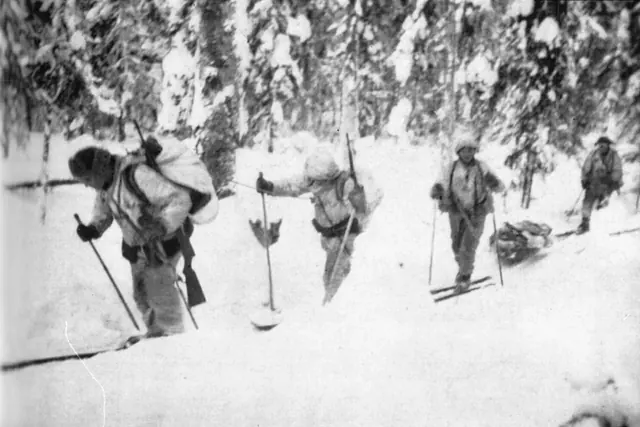 Mustavalkoinen sotilasvalokuva hiihtopartiosta kulkemassa lumisessa metsässä varusteet selässään.